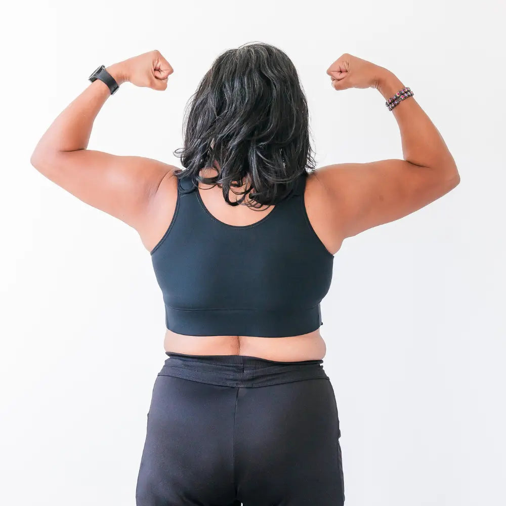 A woman wearing a black resilience bra and leggings, flexing their muscles on a white background